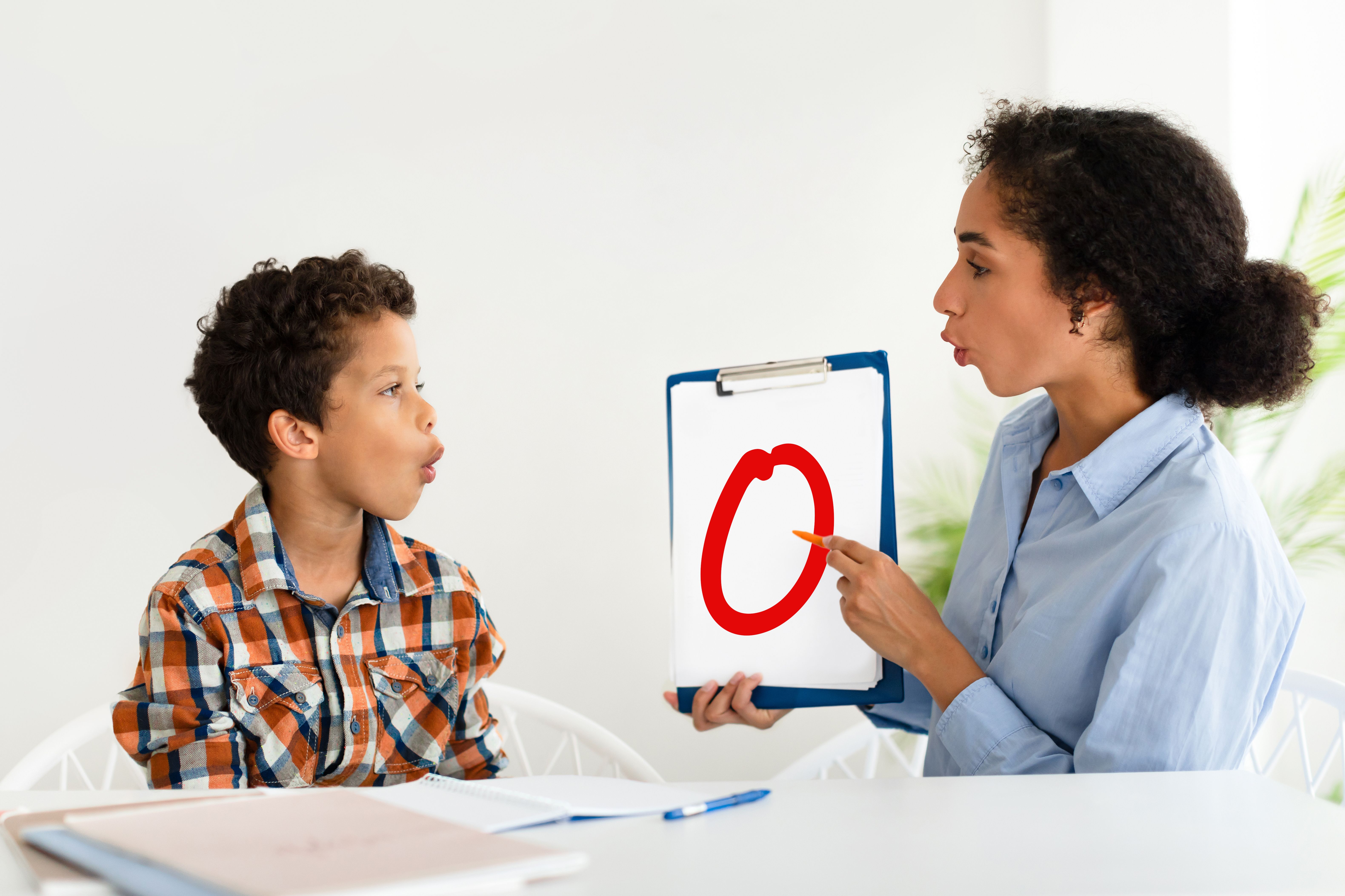 little boy sitting at table miming 'O' as he looks at a picture the Speech Pathologist is holding with the letter 'O' drawn on it