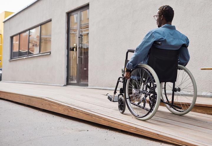 Man in wheelchair going up ramp towards front door of a building