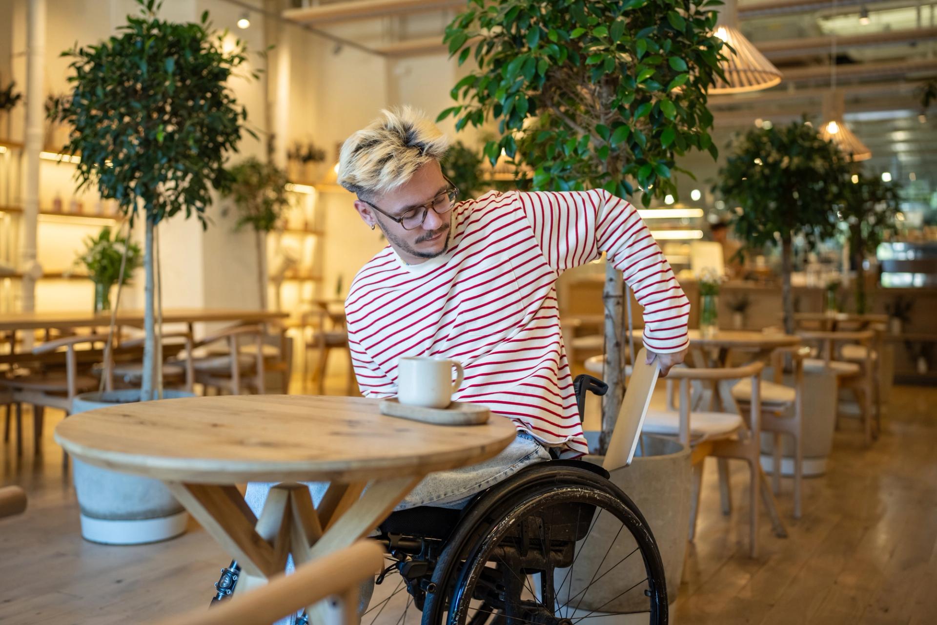 Man in wheelchair sitting at a table in a cafe with a cup of coffee, pulling out an exercise book