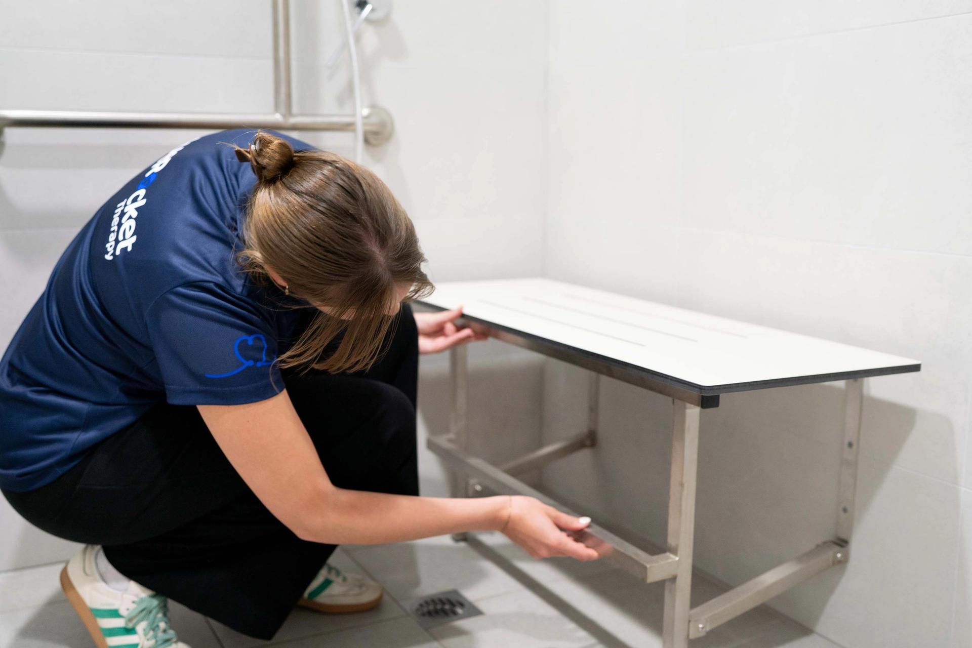 BlueRocket clinician crouched over to adjust a fold out bench in the bathroom