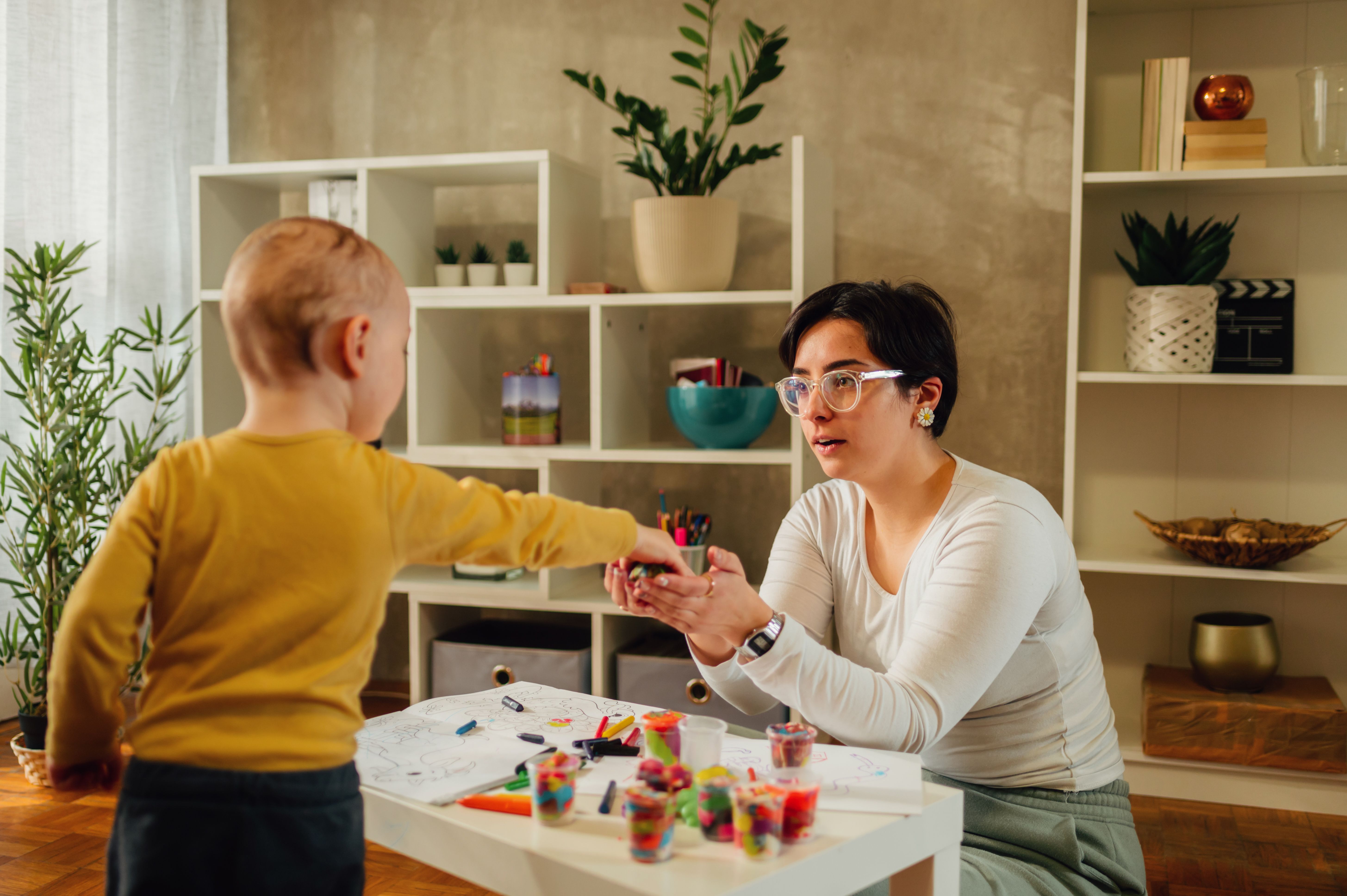 woman sitting at table in a house with young boy handing her playdough