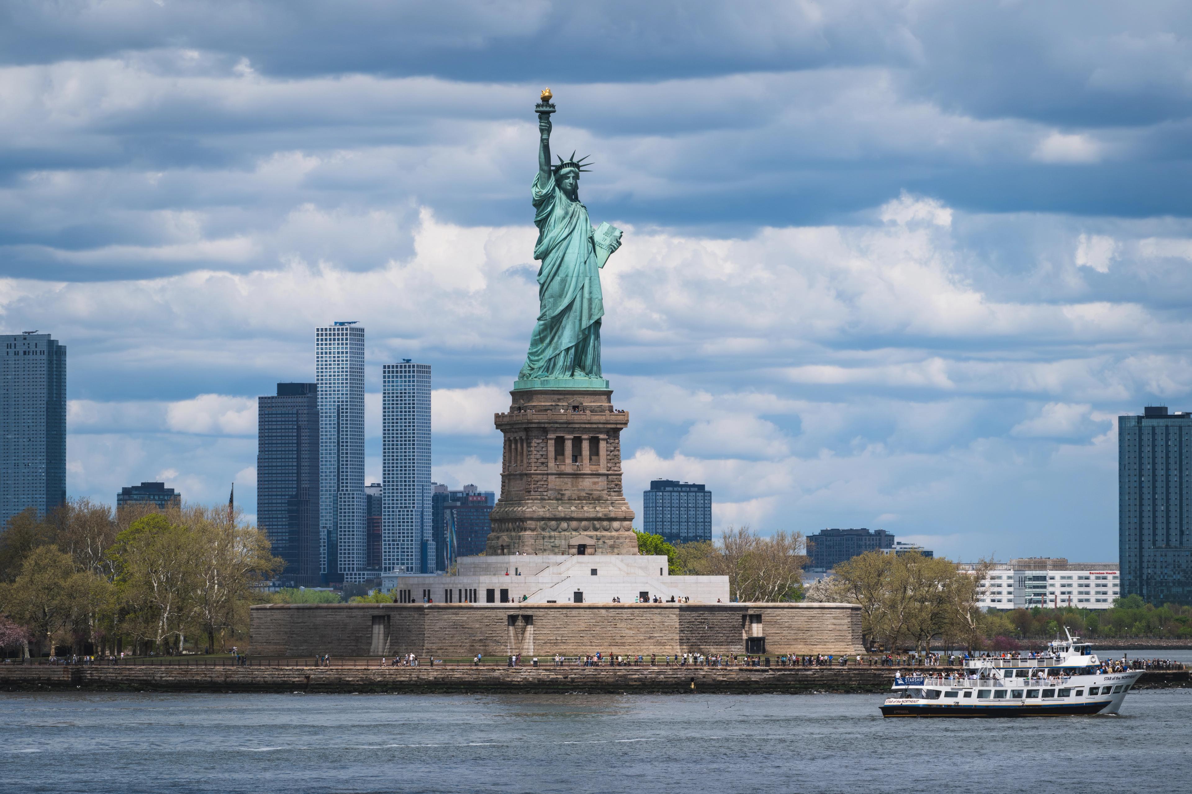 The Statue of Liberty on Liberty Island; a sightseeing boat passes in the foreground with the New Jersey skyline behind.