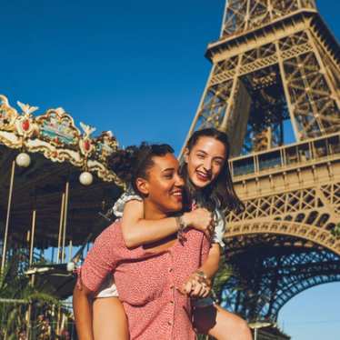 Zwei junge Frauen lachen vor dem Eiffelturm und einem nostalgischen Karussell – erleben Sie den Sommer in Paris mit günstigen Eurostar Zugtickets.