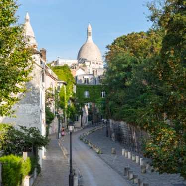 Un jour d'été radieux à la basilique du Sacré-Cœur à Paris, dont les dômes blancs brillent sous la lumière du soleil. Une végétation luxuriante entoure les marches menant à la basilique, où les visiteurs se détendent sur le flanc de la colline herbeuse, profitant d'une vue panoramique sur la ville en contrebas.