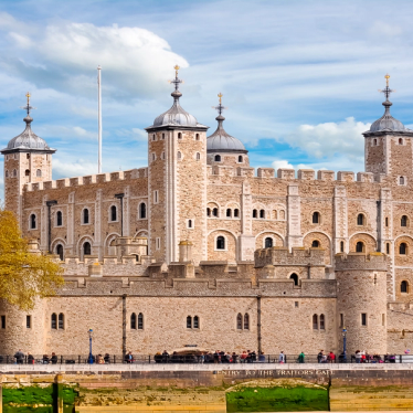 Uitzicht op de Tower of London, met de iconische stenen muren, torens en de beroemde White Tower in het midden. Het historische bouwwerk staat tegen een heldere hemel, met toeristen die het terrein verkennen en de rivier de Theems zichtbaar op de achtergrond.