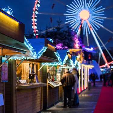 View from below of the Grand Roue in Brussels, beautifully lit up in the background. To the left, a line of Christmas market huts with roofs adorned with twinkling Christmas lights adds to the festive charm. Experience the magic of the Brussels Christmas market with Eurostar.