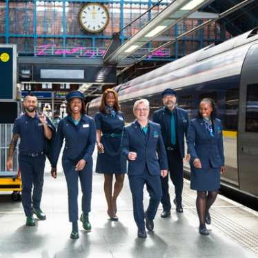 Eine Gruppe uniformierter Zugmitarbeitender geht gemeinsam auf einem Bahnsteig neben einem Zug und lächelt.