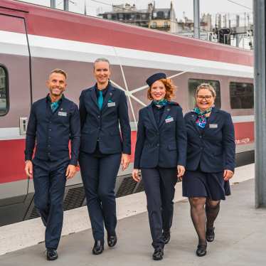 Vier uniformde treinmedewerkers lopen samen over een perron naast een trein en glimlachen.