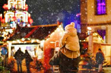 Ein Vater, der sein Kind auf den Schultern trägt, vor einem verschneiten Hintergrund mit leuchtenden Weihnachtsmarktlichtern auf einem der schönsten Weihnachtsmärkte in Europa. 