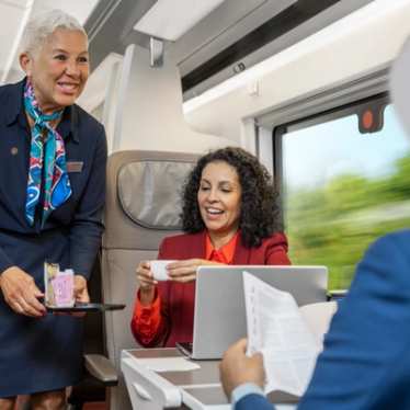 A staff member serves a drink to a passenger working at her seat on the train.