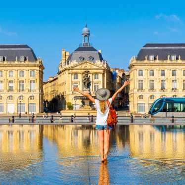 Happy woman tourist with arms raised in Place de la Bourse square with water mirror, Bordeaux city landscape.