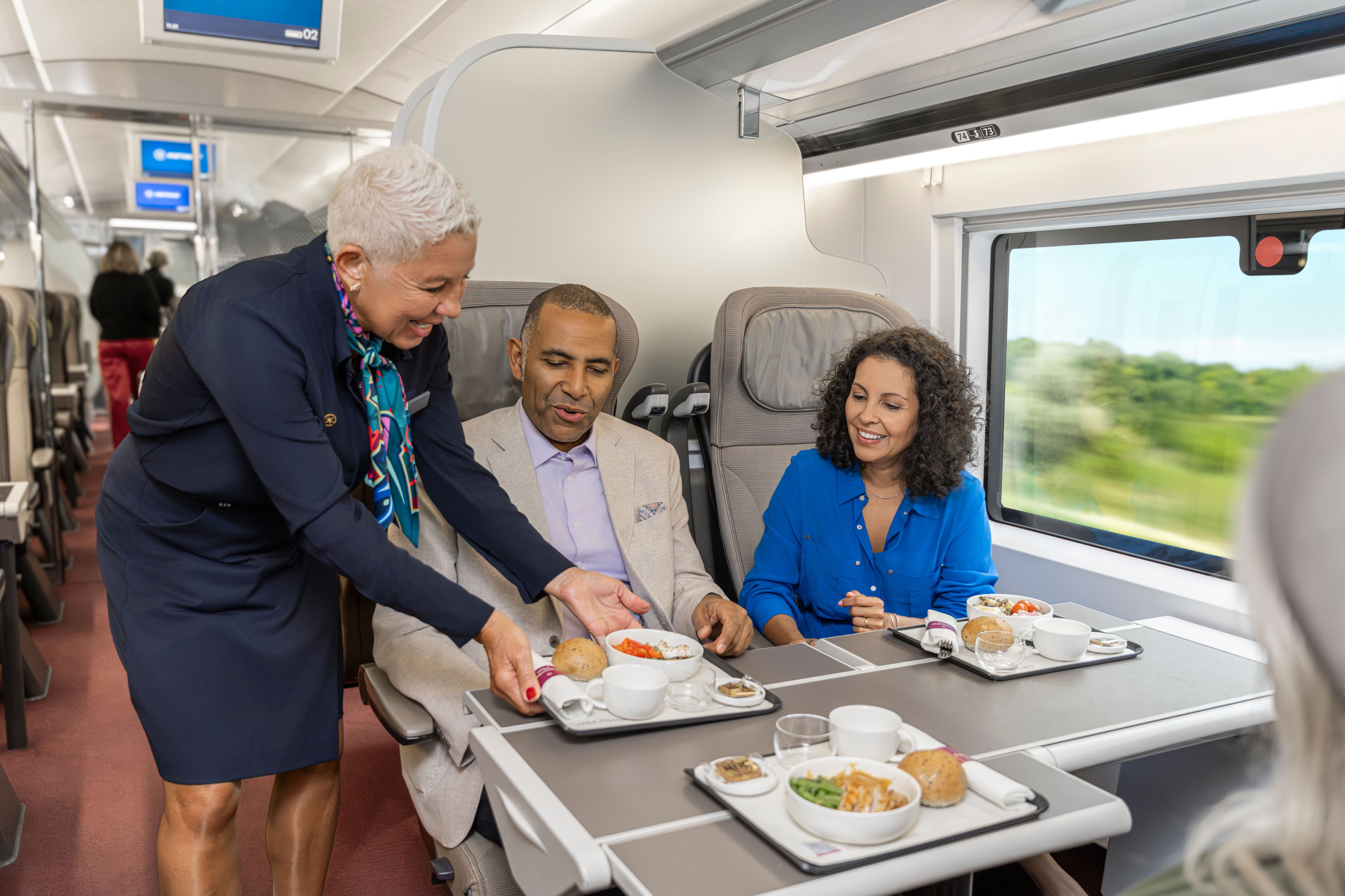 A staff member serves meals to two passengers seated together on the train.