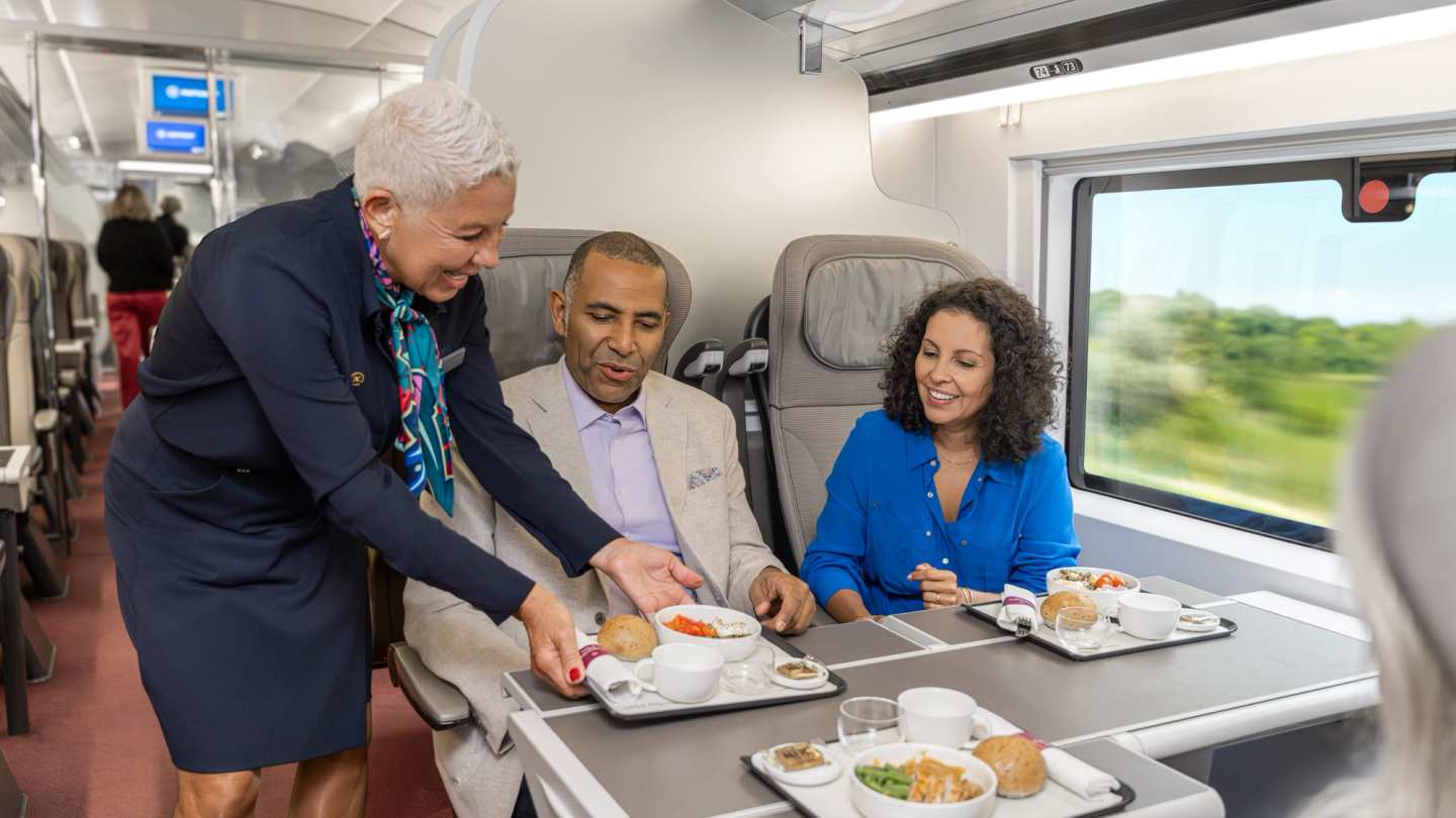 A staff member serves meals to two passengers seated together on the train.