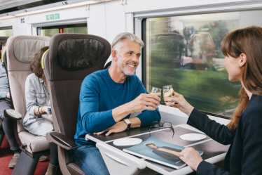 Two travellers, a man and a woman, sitting opposite onboard a train, raising a toast.