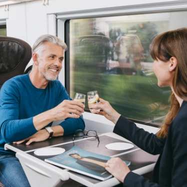 Two travellers, a man and a woman, sitting opposite onboard a train, raising a toast.