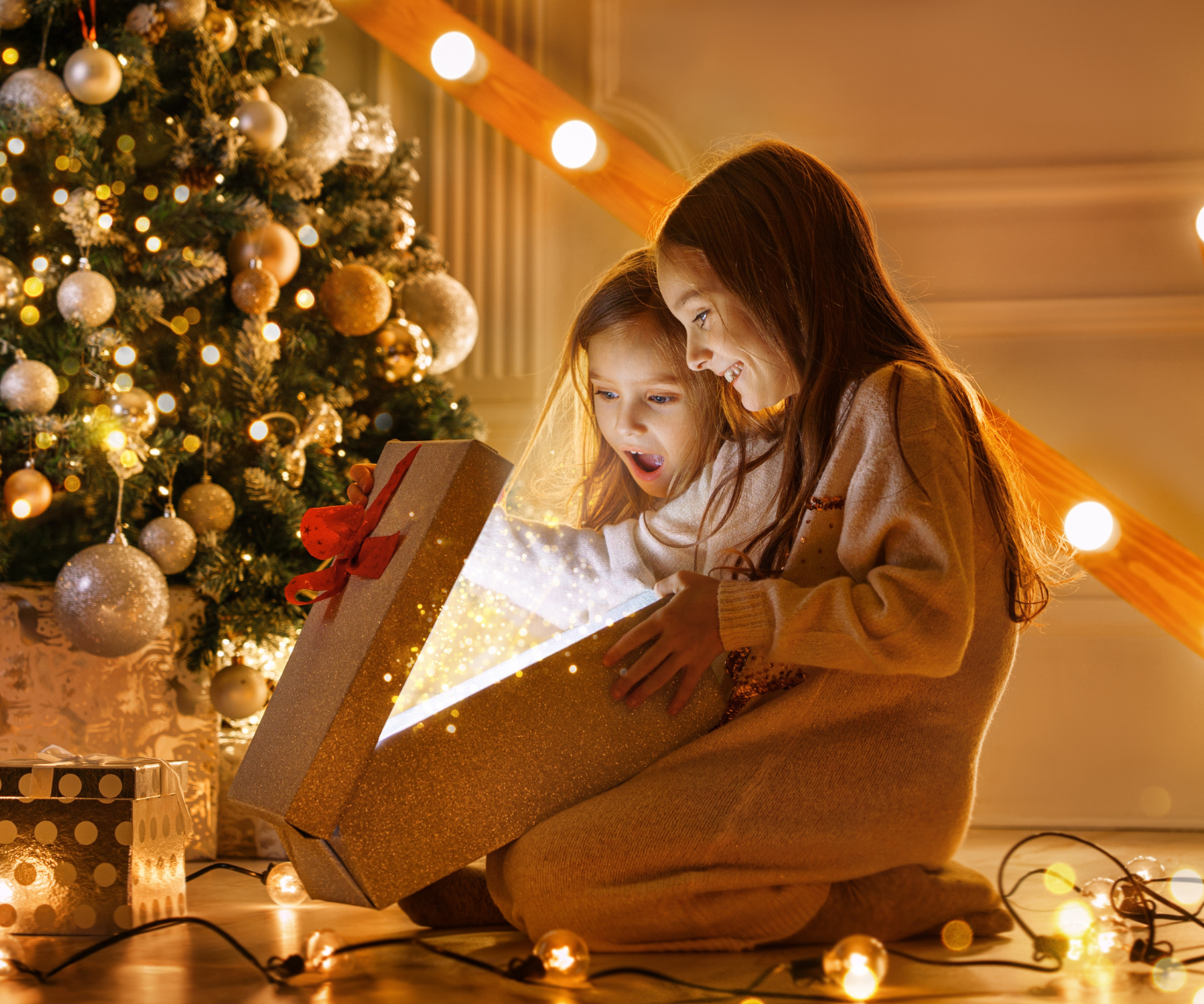 Two little girls opening a Christmas present in front of a softly golden lit tree