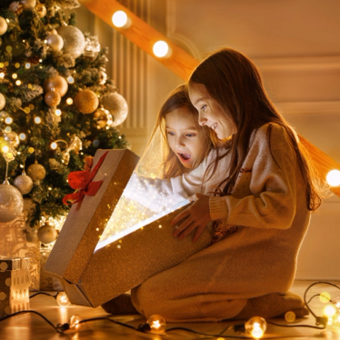 Two little girls opening a Christmas present in front of a softly golden lit tree