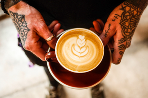 A man with hand tattoos holding a latte displaying the art on top.