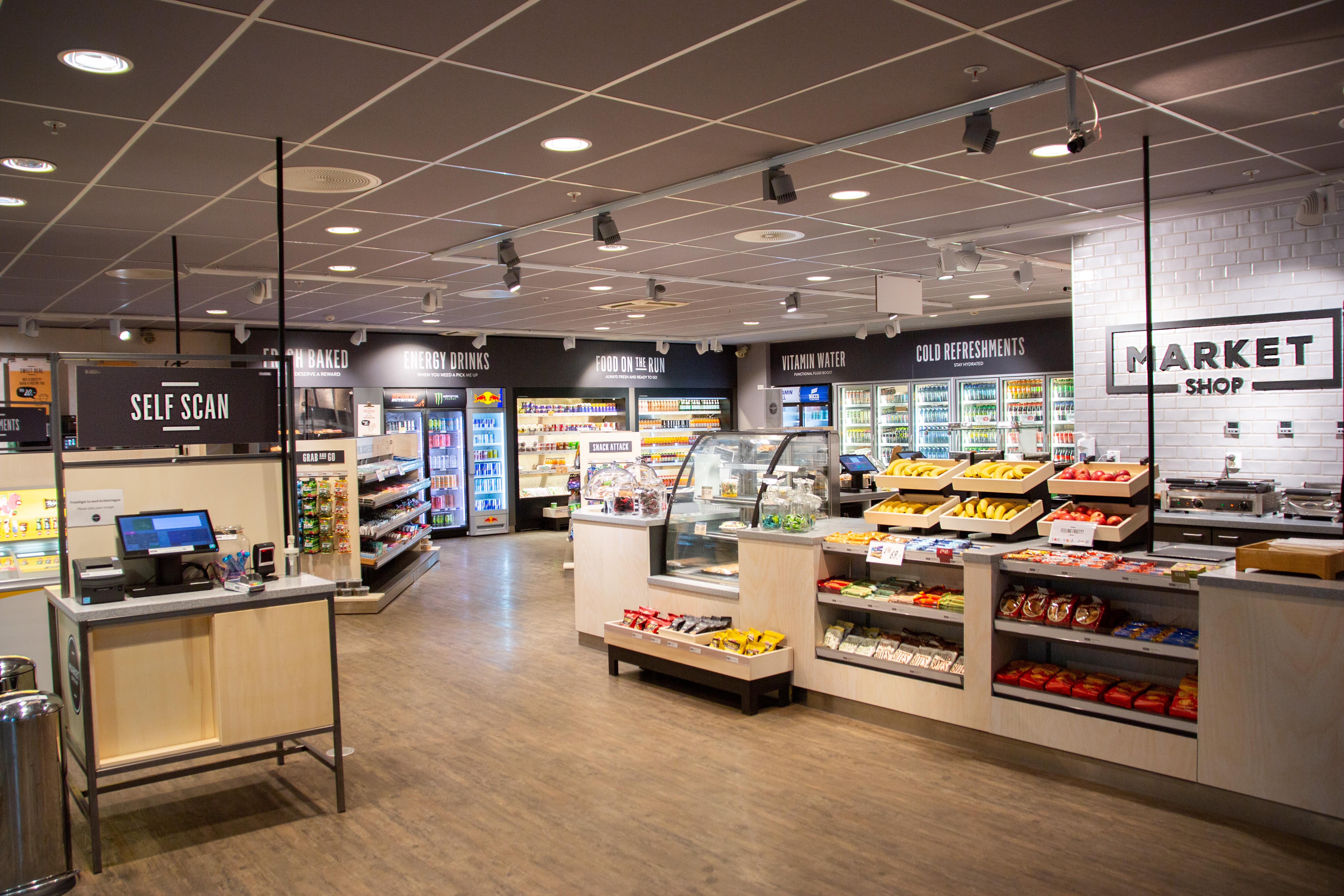 Interiør i Market shop kiosk. Disk med varer, hyller med varer og self scan-løsning.
