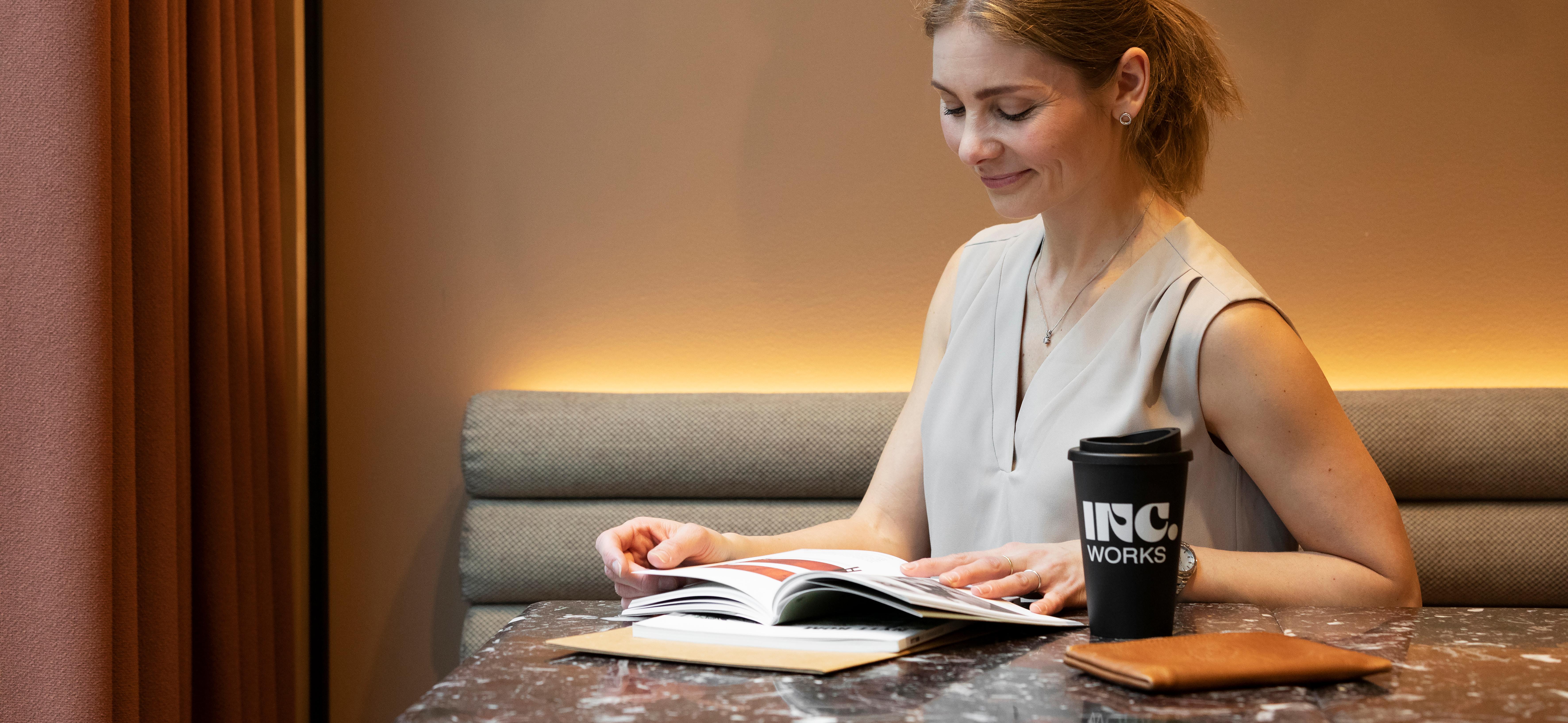 Woman sits at a table and reads, and has a coffee cup next to her with INC. logo on it