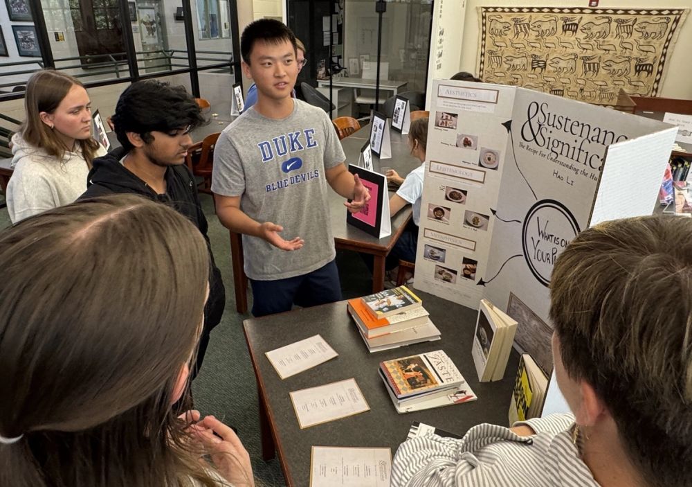 A student presents a tri-fold research display board titled "Sustenance & Significance" to classmates during a research project showcase.