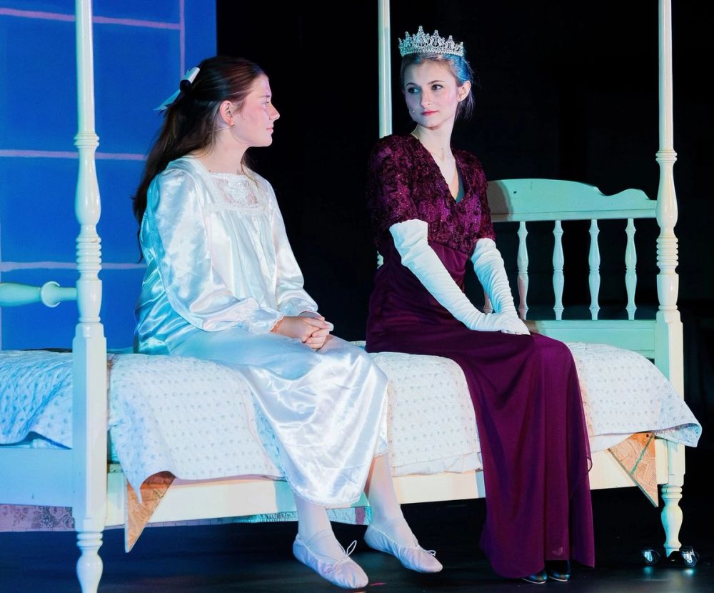 Two student actors perform a scene in period costume on stage, one seated on a bed.
