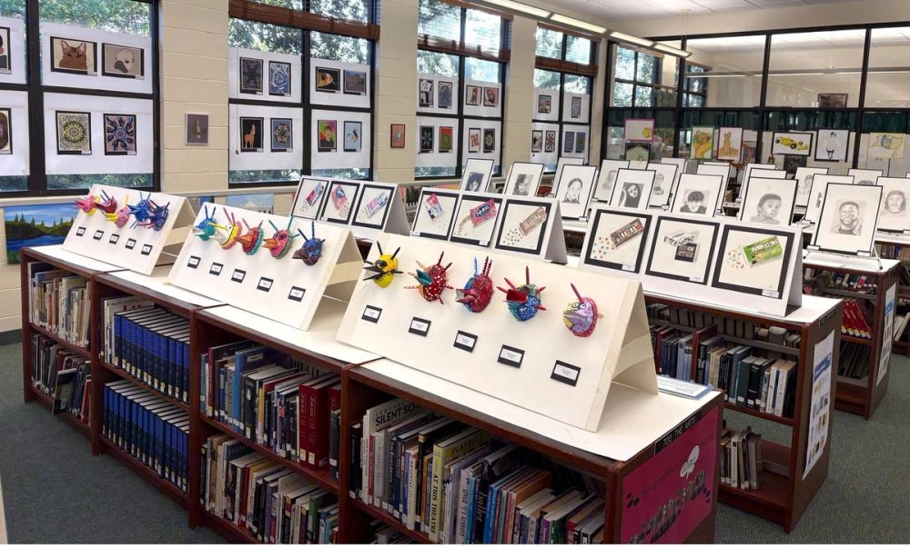 A library display of student artwork and origami on stands beside framed pieces.