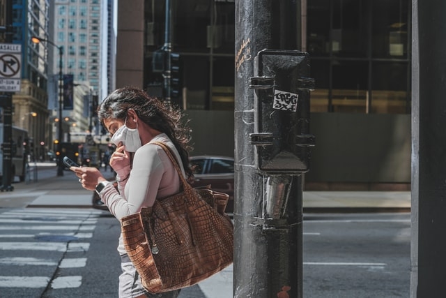 A woman in an N95 mask walks down a downtown street looking at her phone. 