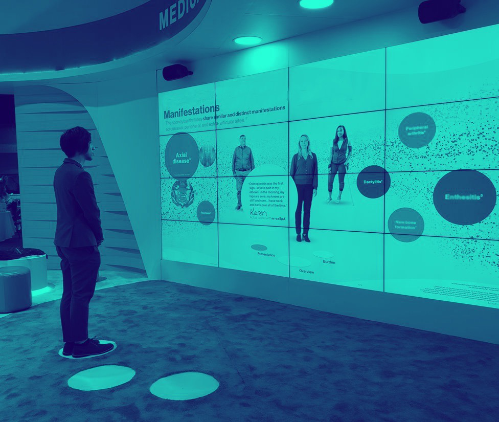 Duotone teal photo of a person at a conference standing in front of an interactive wall, that reacts based on their position on the floor. The display shows multiple people and bubbles of information. 