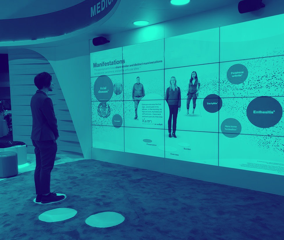 Duotone teal photo of a person at a conference standing in front of an interactive wall, that reacts based on their position on the floor. The display shows multiple people and bubbles of information.