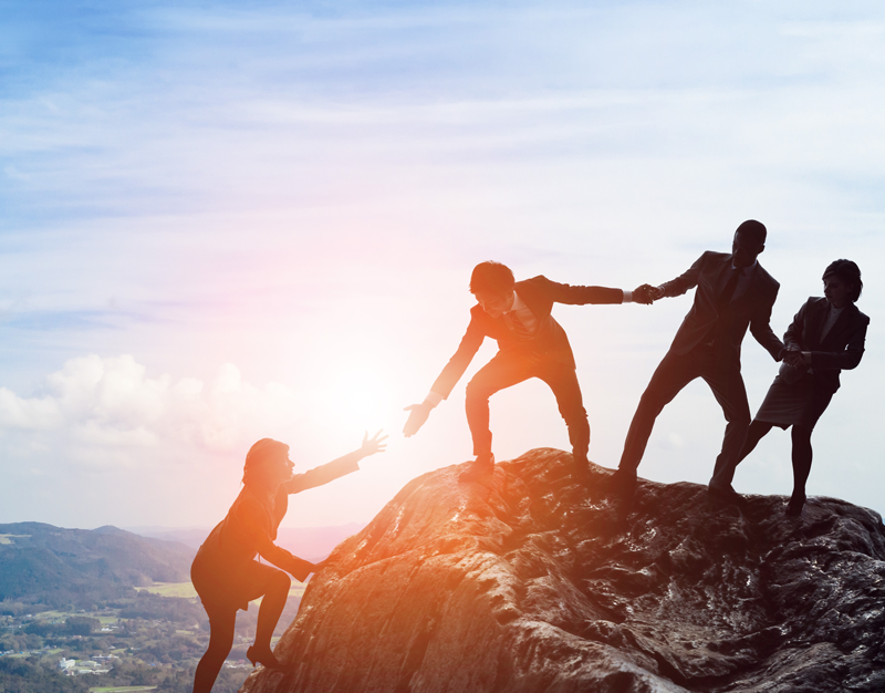 People in business attire help each other scale a summit while the sun makes them silhouettes. 