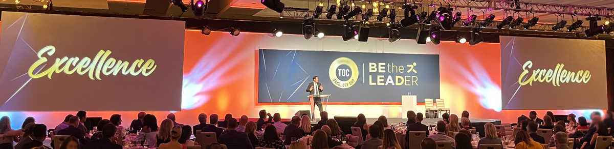 Conference stage with speaker, and 3 screens saying "excellence" and Be the leader". Fuschia and orange lighting.