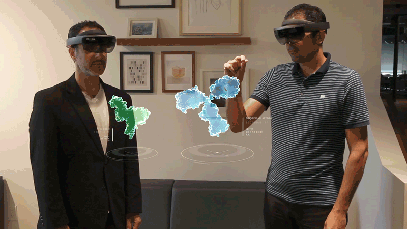 Two men wearing HoloLens 2 headsets manipulate molecules together in mid-air in front of them. We're seeing what they see.