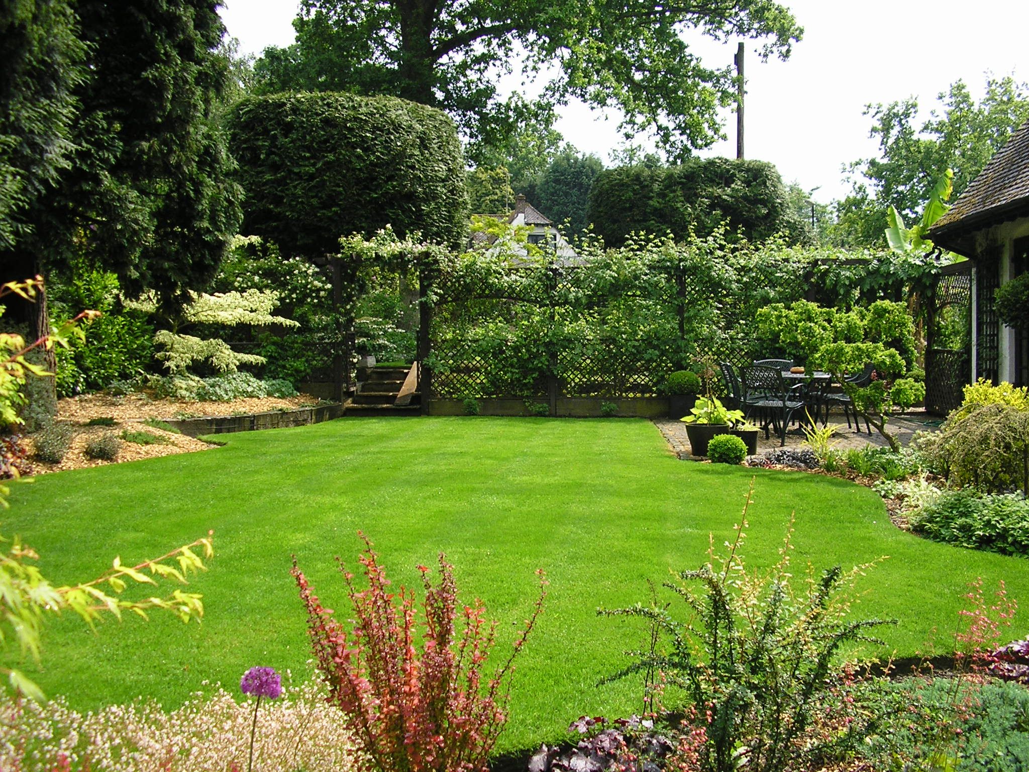 A well-kept garden with a lush green lawn, surrounding shrubs, and a patio seating area in the background.