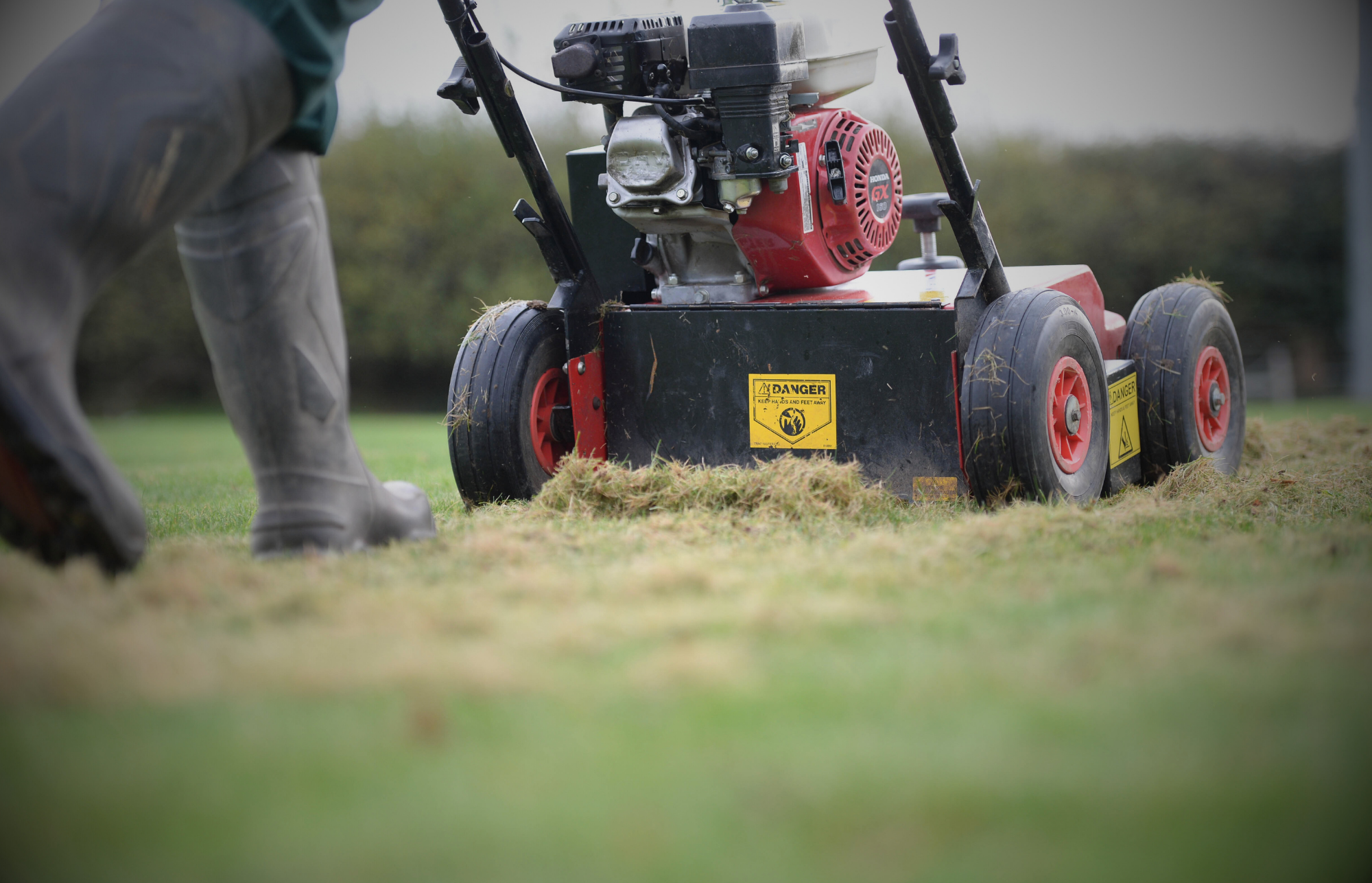 Lawn scarification machine removing thatch from grass, with a person in rubber boots walking beside it. Warning labels are visible on the machine.