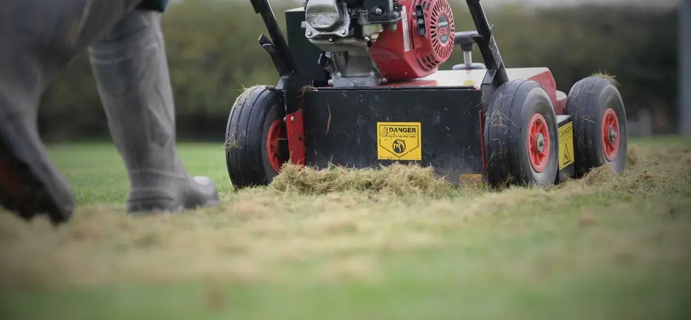 Lawn scarification machine removing thatch from grass, with a person in rubber boots walking beside it. Warning labels are visible on the machine.
