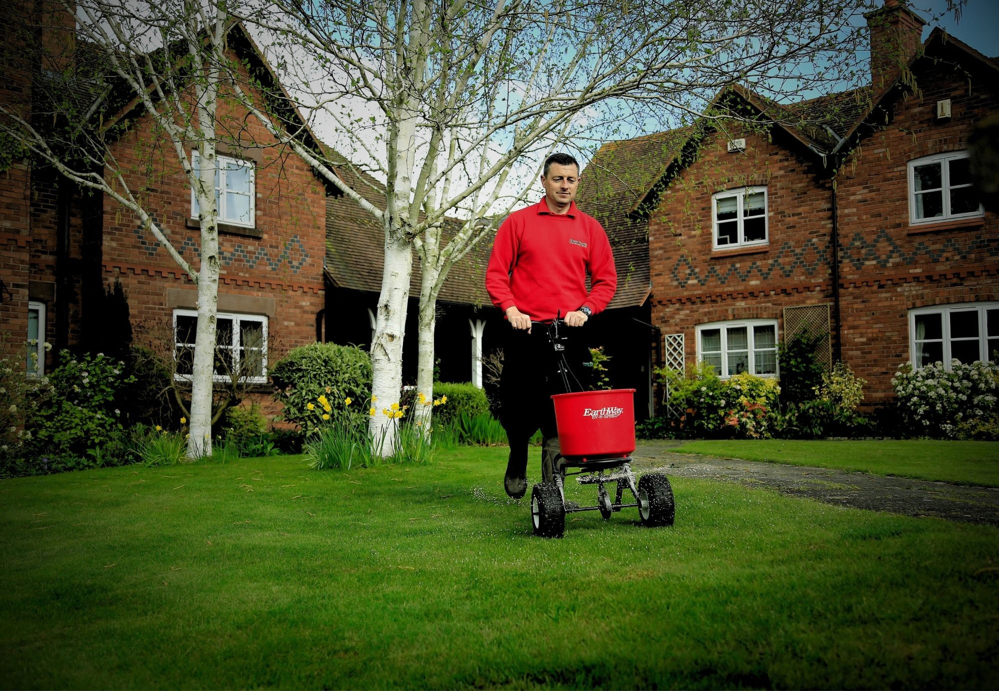 GreenThumb lawn care specialist applying professional lawn treatment with a spreader in front of a traditional brick house and garden.