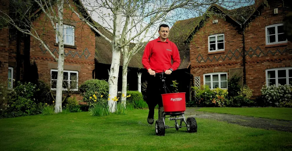 GreenThumb lawn care specialist applying professional lawn treatment with a spreader in front of a traditional brick house and garden.
