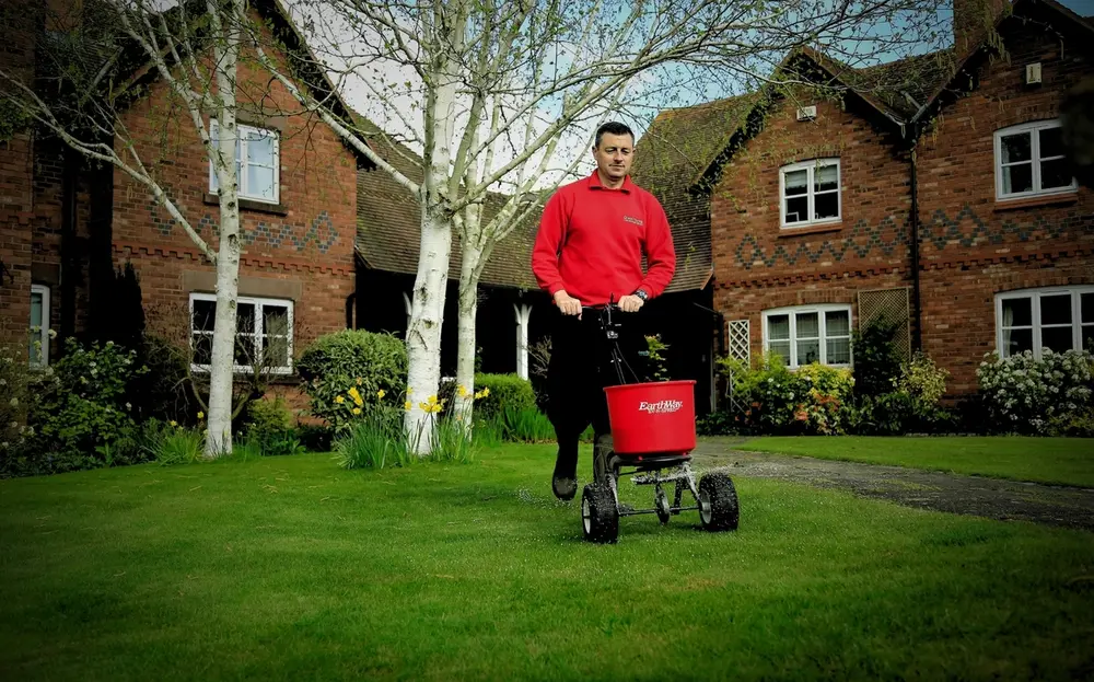 GreenThumb lawn care specialist applying professional lawn treatment with a spreader in front of a traditional brick house and garden.