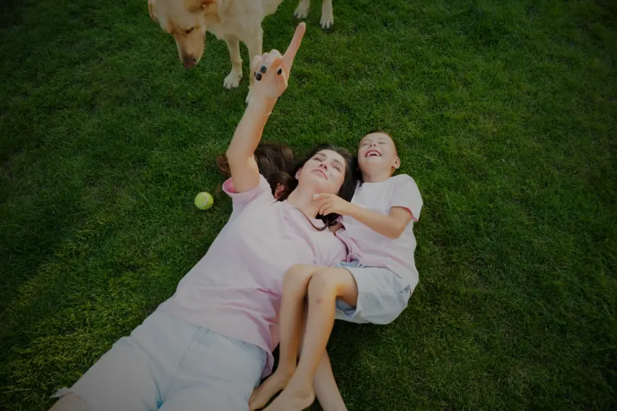 A smiling mother and child lying on a healthy lawn with their dog nearby, enjoying quality time outdoors on lush green grass.