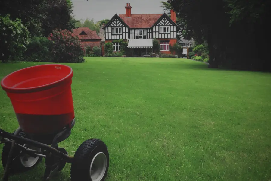 A lush green lawn being treated with a red push spreader, with a large Tudor-style house and garden in the background.