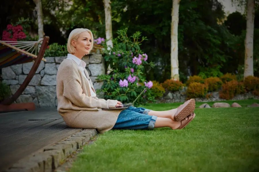A woman sits on a garden deck with her laptop, surrounded by blooming plants and lush lawn—enjoying a peaceful, outdoor workspace.