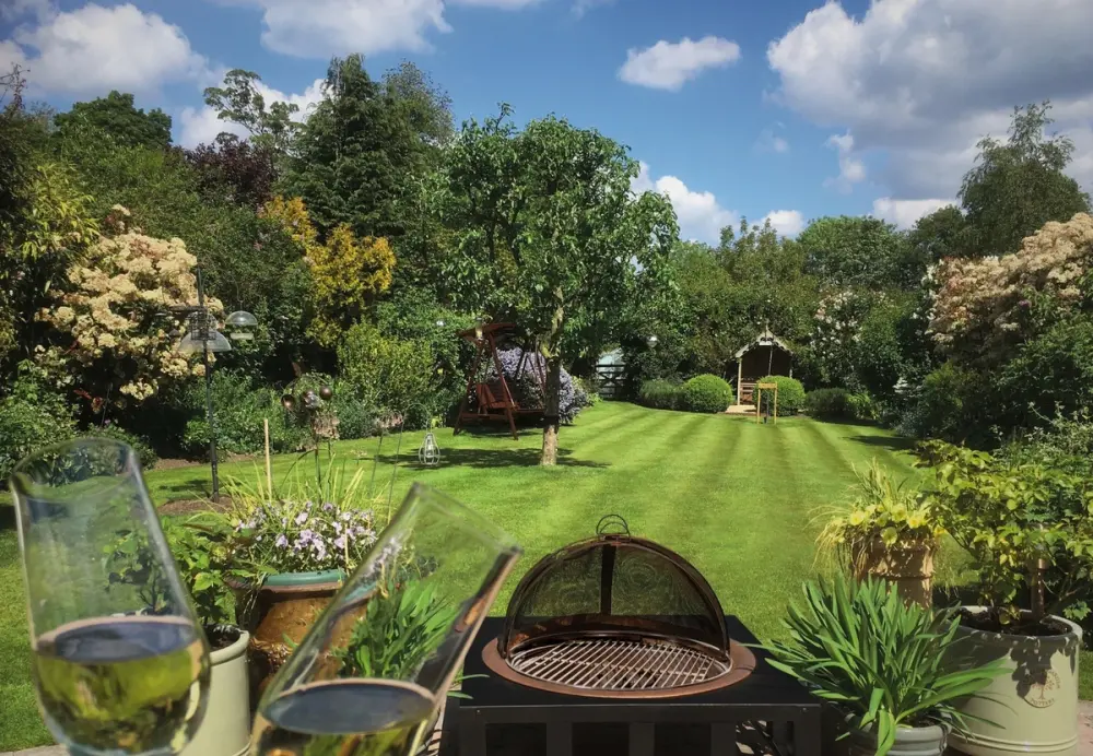 Two glasses raised in front of a lush striped lawn with garden seating under a blue summer sky.