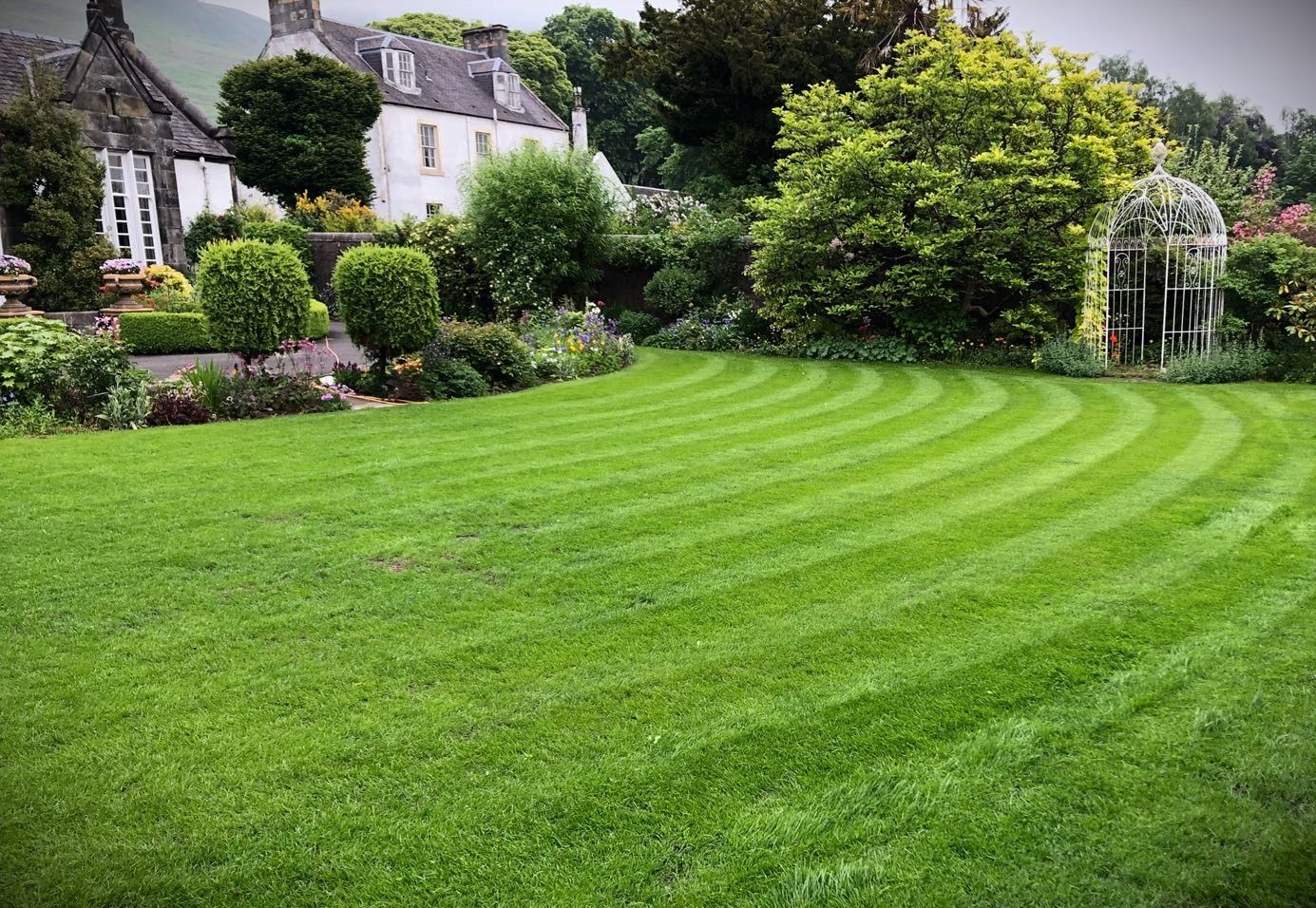 A beautifully striped, well-maintained lawn in front of a traditional cottage garden with neatly trimmed hedges, trees, and a white birdcage-style garden feature.