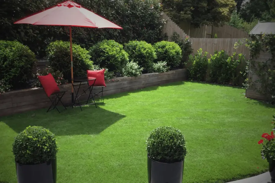 A neat, vibrant green lawn with patio seating and a red umbrella, bordered by raised flower beds and lush garden planting.