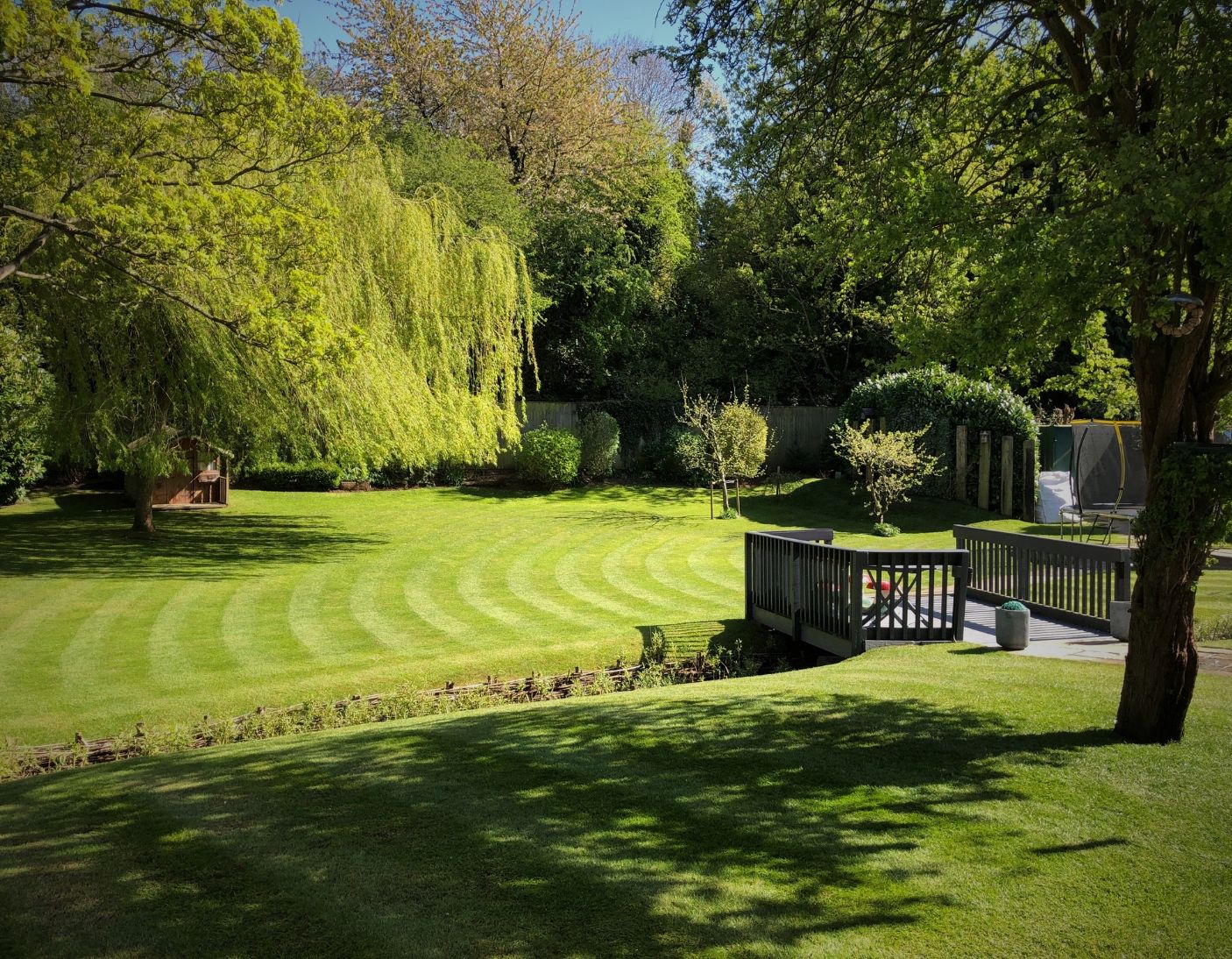 A neatly striped lawn in a sunlit garden with mature trees, manicured hedges, and a wooden deck area overlooking the grass.