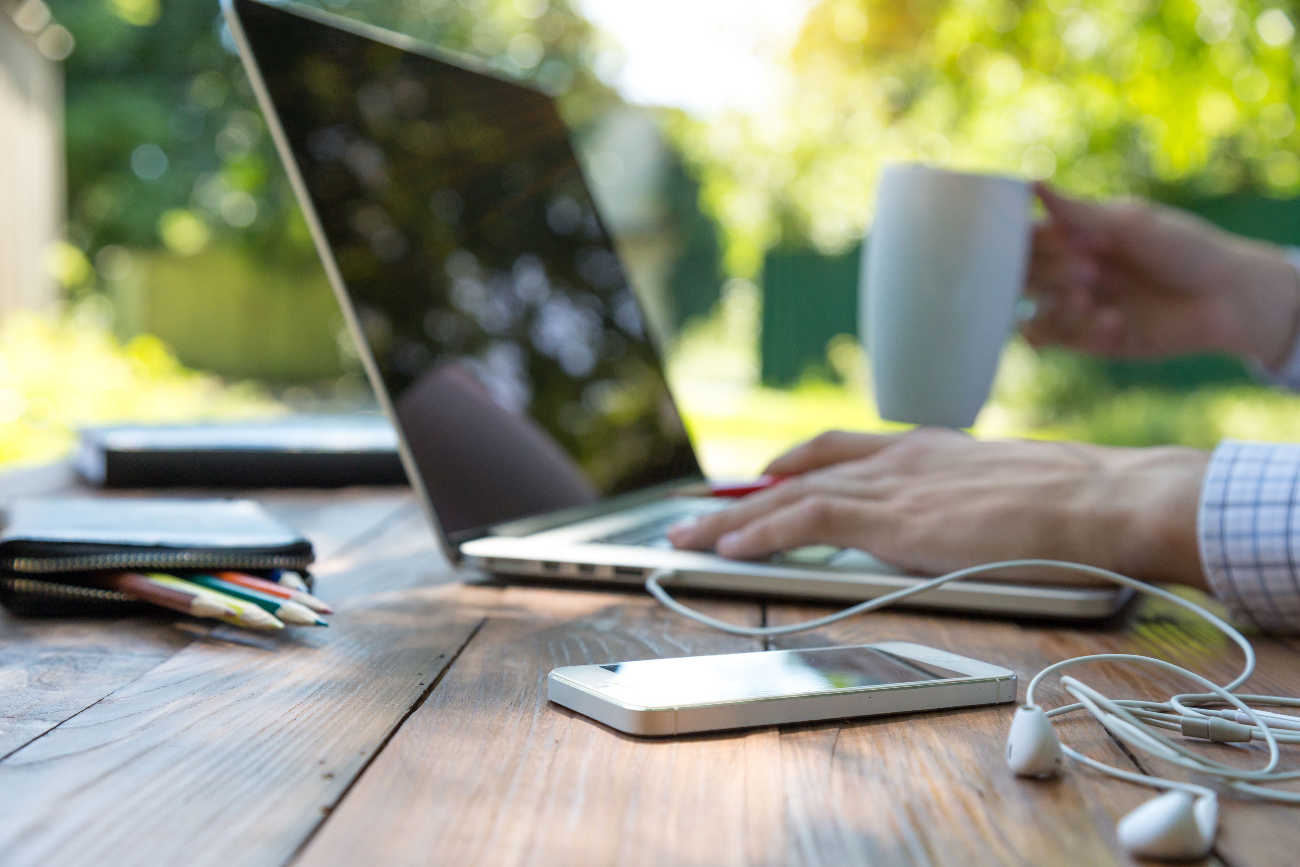 Person using a laptop outdoors with a coffee, phone, and notebook on the table.