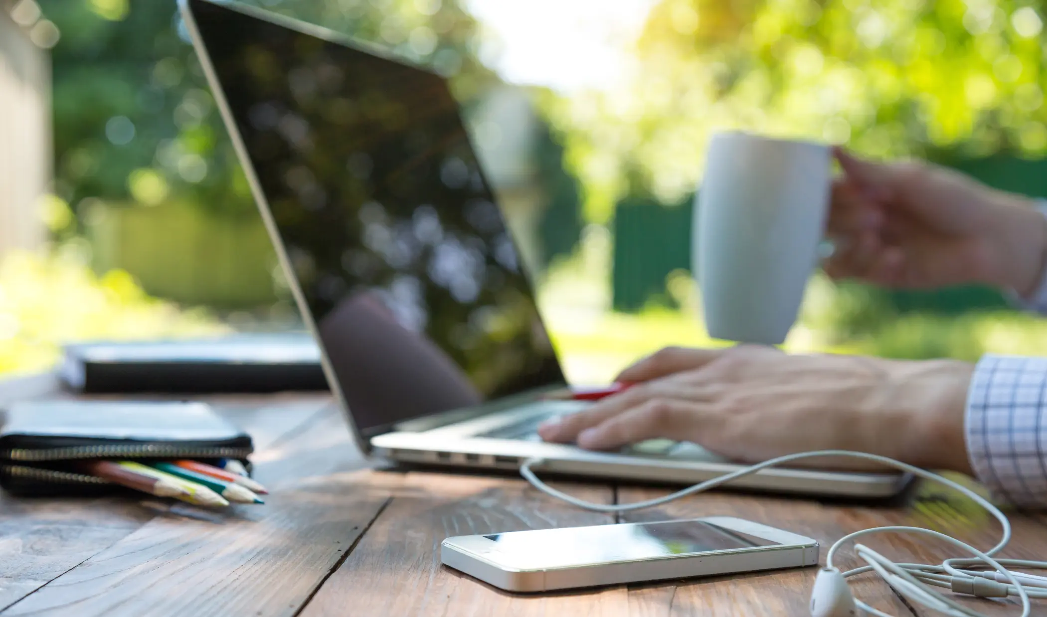 Person using a laptop outdoors with a coffee, phone, and notebook on the table.