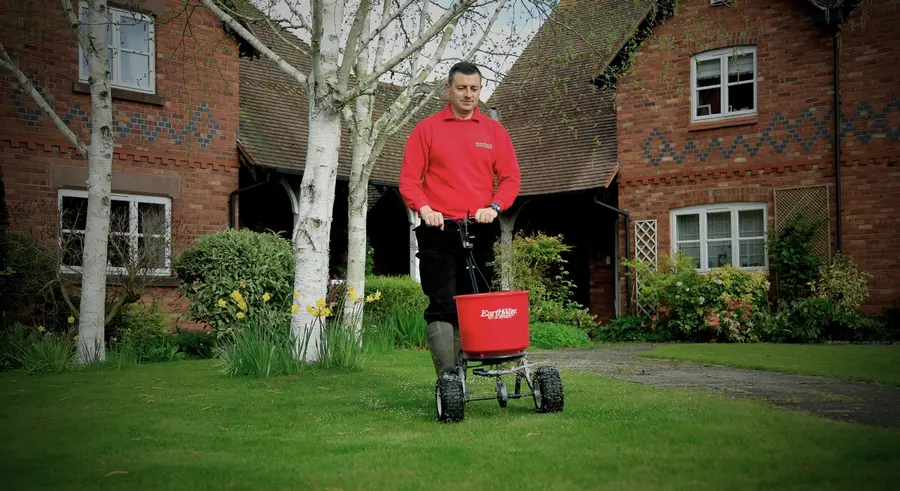 GreenThumb lawn specialist applying treatment using a fertiliser spreader outside a residential home.
