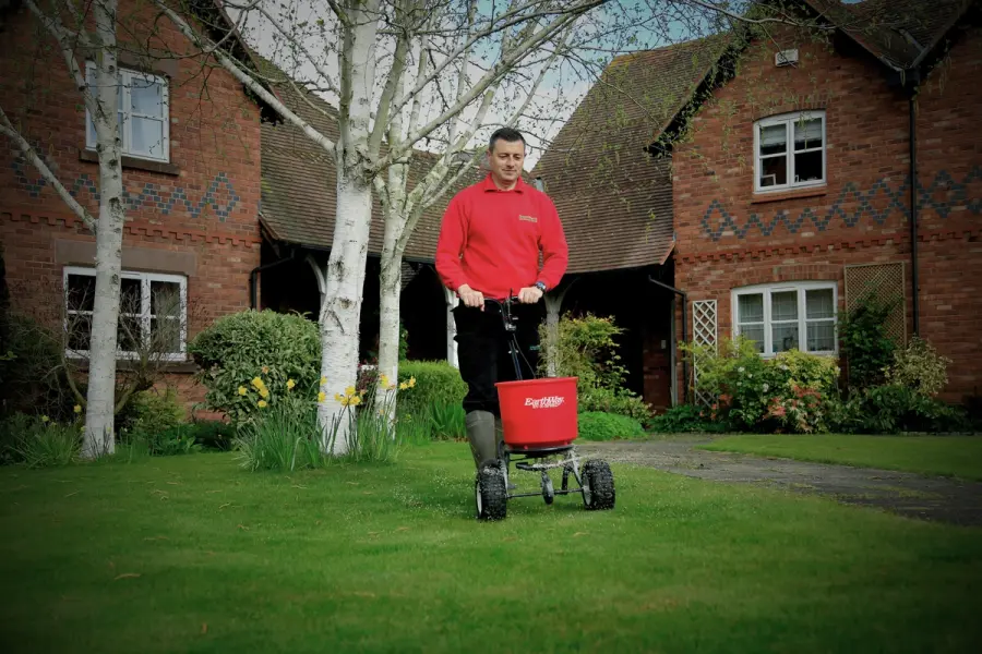 GreenThumb lawn expert applying treatment outside a brick home using a broadcast spreader, promoting healthy green grass.